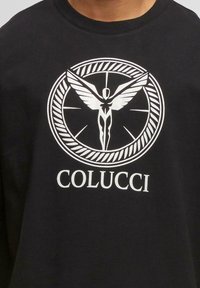 Schwarzes Hemd mit einem weißen runden Logo, das eine geflügelte menschliche Figur zeigt, und dem Text "COLUCCI" unter dem Design.