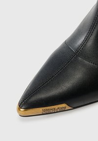 Black leather pointed-toe ankle boot featuring a textured surface and a gold accent on the heel with "VERSACE JEANS COUTURE" branding.