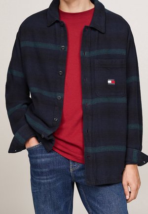 Personne portant une chemise à carreaux bleu foncé avec poche poitrine, par-dessus un t-shirt rouge et un jean bleu, main dans la poche.