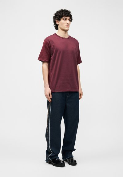 Νέος άνδρας με κατσαρά μαλλιά, που φοράει άνετο μπορντό T-shirt, σκούρα τζιν με ραφή στο πλάι και μαύρα γυαλιστερά παπούτσια φόρμας, στέκεται μπροστά σε απλό φόντο.