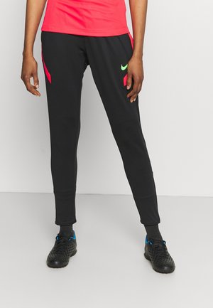 Person trägt schwarze Sporthose mit neongrünem Nike-Logo, schwarze Fußballschuhe und ein rotes Shirt vor weißem Hintergrund.