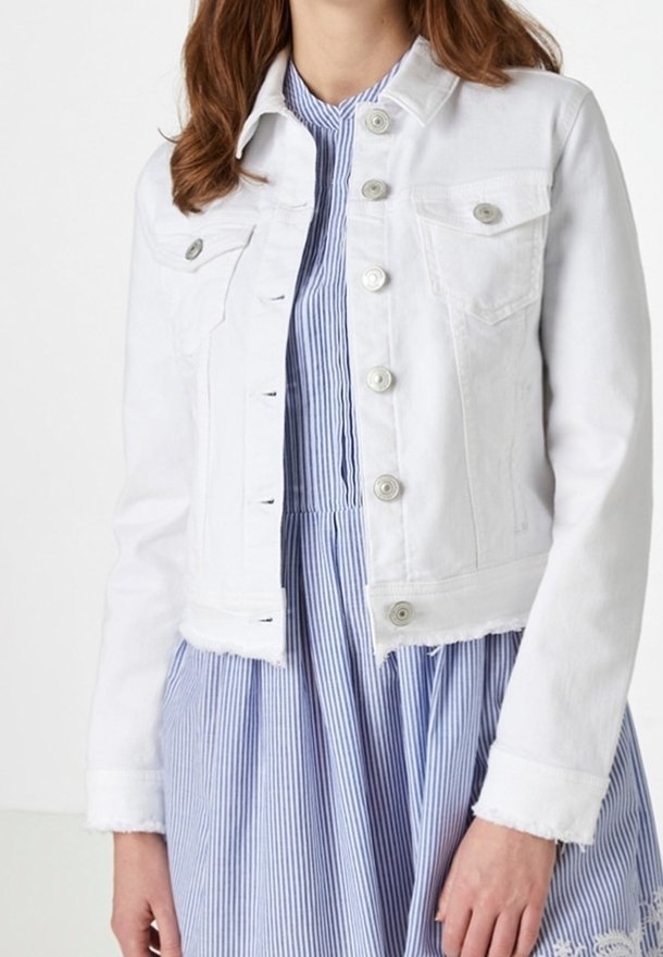 Bílá džínová bunda s knoflíky přehozená přes modro-bílé šaty s vertikálním pruhem a krajkovým lemem, které nosí stojící osoba.