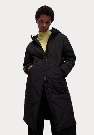 Manteau en duvet noir avec capuche, motif matelassé en losanges, fermeture éclair et poches latérales. Porté sur un col roulé jaune clair.