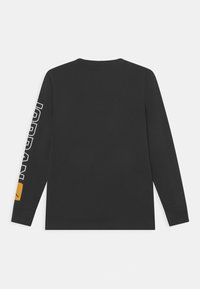 Pitkähihaisen mustan t-paidan materiaali on puuvillaa, ja siinä on valkoinen "JORDAN" -teksti vasemmassa hihassa sekä keltainen logoprintti alareunassa.