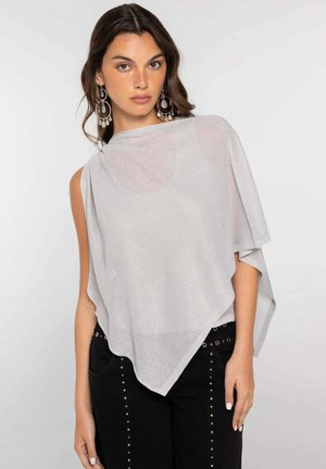Mujer con cabello largo y oscuro lleva un poncho asimétrico gris claro y pendientes grandes de plata, combinados con pantalones negros con tachuelas.