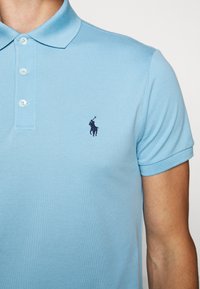 Polo bleu clair à manches courtes avec trois boutons blancs et un logo brodé foncé représentant un cheval et un cavalier sur la poitrine gauche.