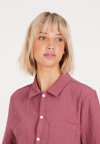 Camisa color burdeos con superficie texturizada, frente con botones y un solo bolsillo en el pecho. Presenta un cuello clásico y una tela ligera.