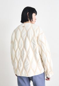 Quilted cream jacket with beige diamond patterns, round neckline, and button details at the back, paired with light blue pants.