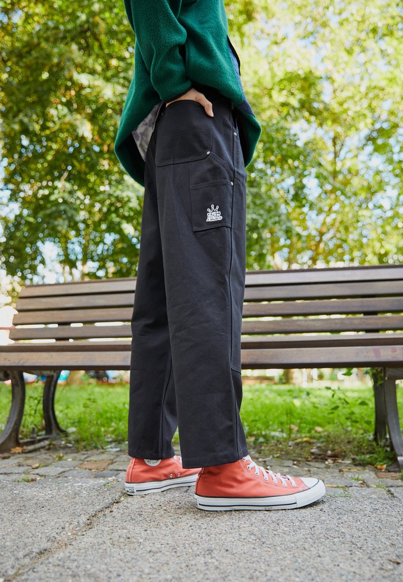 Svarta bomullbyxor med en avslappnad passform, med sidofickor och en liten vit logotyp. Pairade med orange högskoor.