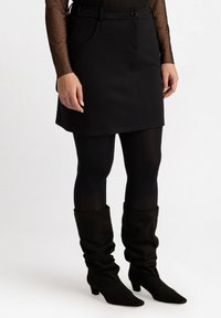 Svart denimkjol med fickor, knäppning med knappar och bältesögglor, tillsammans med svarta täckande strumpbyxor och slappa svarta mocka ankelstövlar.