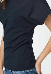 Femme portant un haut noir froncé à manches courtes associé à un jean en denim bleu, la main posée sur la hanche, le torse visible sur un fond uni.