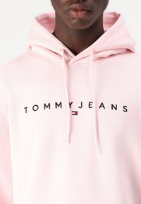 Hanorac roz din material moale, cu glugă ajustabilă și cuvintele "TOMMYJEANS" scrise cu negru, având un mic detaliu cu logo în design.
