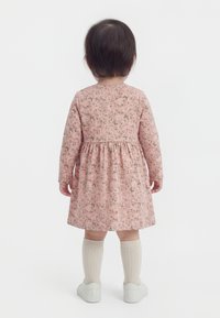 Niño pequeño con cabello corto y oscuro, vestido con un vestido largo de manga larga y flores rosas, calcetas crema hasta la rodilla y zapatos blancos, de pie y de espaldas.