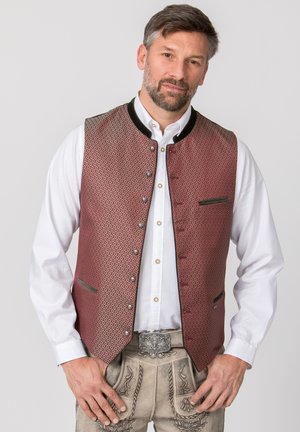 Bärtiger Mann, der eine gemusterte rote Weste über einem weißen Hemd mit bestickten beigen Lederhosen und einer dekorativen Gürtelschnalle trägt.