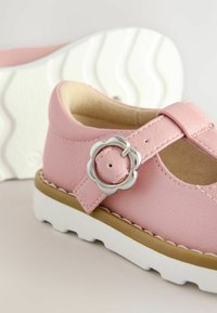 Rosa Lederkinderschuhe mit T-Riemen-Design, silberner Blumen-Schnalle, weißer gewellter Sohle und genähten Details entlang der Kanten.