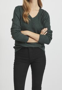 Ciemnozielony, miękki sweter z dzianiny z dekoltem w serek, ściągaczami na mankietach i dole; w połączeniu z czarnymi dopasowanymi dżinsami, ręce skrzyżowane przed sobą.