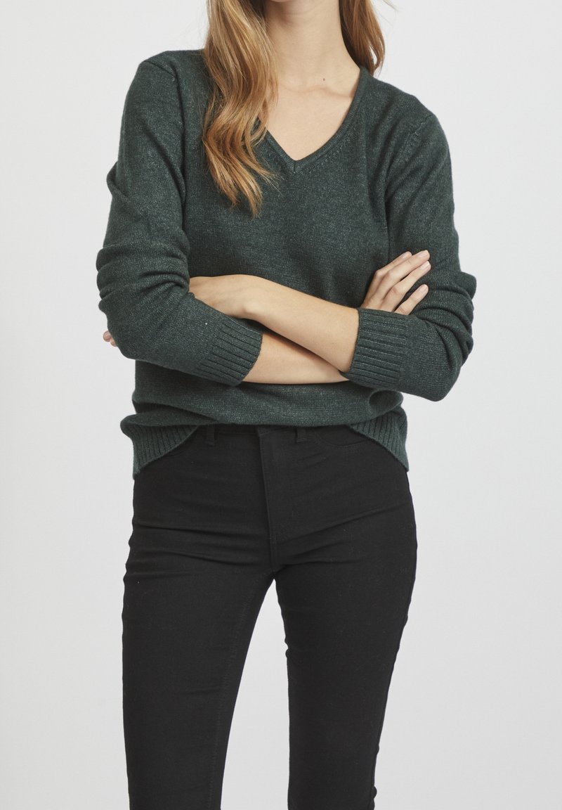 Ciemnozielony, miękki sweter z dzianiny z dekoltem w serek, ściągaczami na mankietach i dole; w połączeniu z czarnymi dopasowanymi dżinsami, ręce skrzyżowane przed sobą.