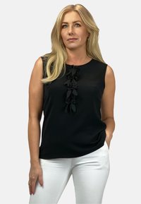 Czarny bezrękawnik z falbaną na środku, zestawiony z białymi spodniami. Gładki materiał i luźny krój.