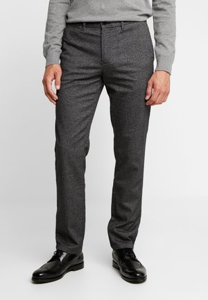 Homme portant un pantalon de costume gris foncé, un pull gris clair et des chaussures en cuir noir, debout sur un fond clair uni.