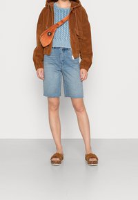 Brun manchesterjacka med dragkedja, ljusblå texturerad stickad topp, ljusa jeansshorts, bruna sandaler med knut och mönstrade sulor. Orange axelremsväska.