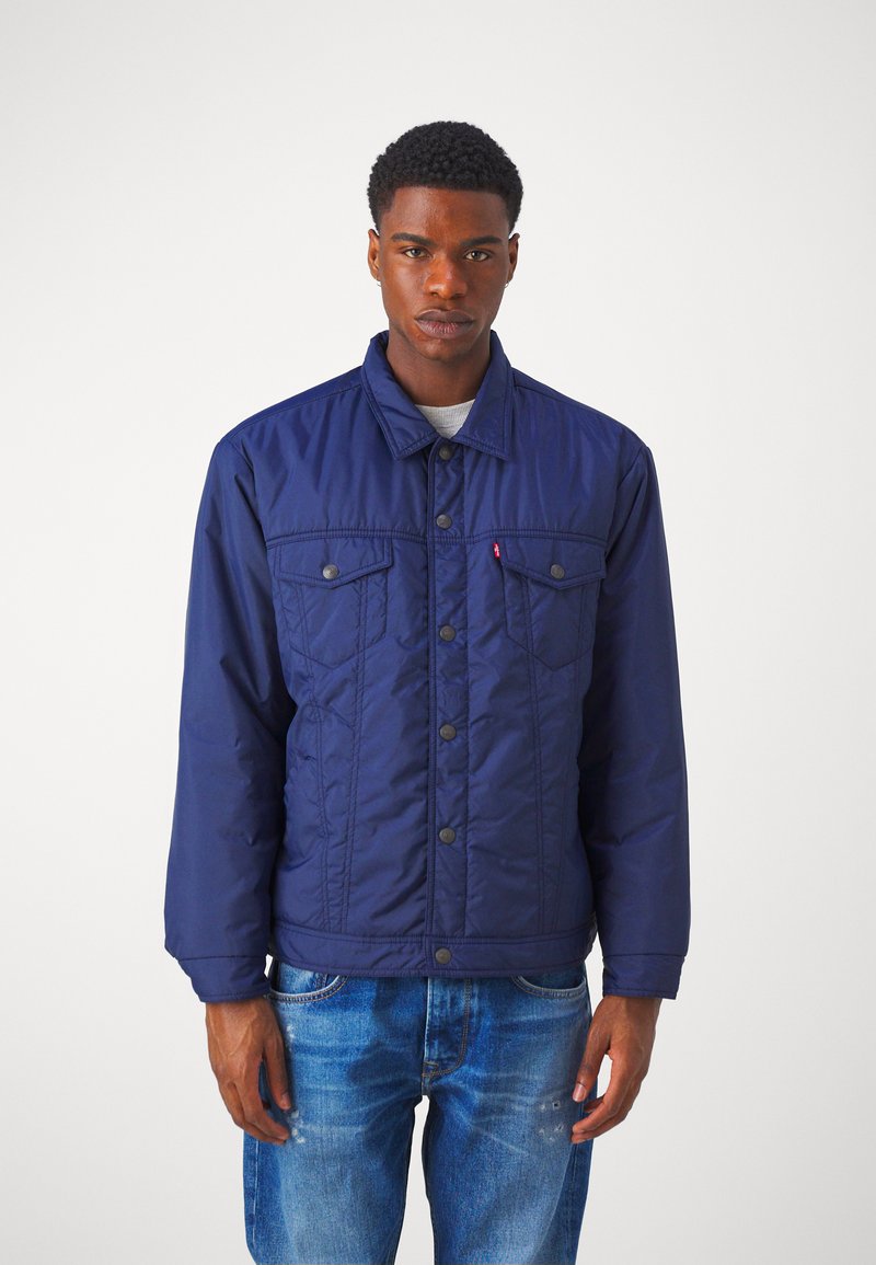 Chaqueta acolchada de color azul marino con cuello tipo camisa, dos bolsillos en el pecho y botones de presión. Tejido texturizado y corte relajado, combinada con jeans de mezclilla claros.