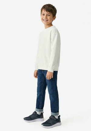 Lächelnder Junge steht, trägt ein weißes Langarmshirt, blaue hochgekrempelte Jeans, weiße Socken und schwarze Sneaker mit Klettverschlüssen.