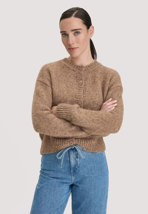Junge Frau mit dunklem, zurückgebundenem Haar trägt einen braunen, geknöpften Strickpullover und blaue Jeans und steht mit verschränkten Armen vor einem einfarbigen Hintergrund.