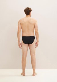 Pantaloni da bagno neri, con un design semplice e una texture liscia, indossati da una persona dalla pelle nuda. Vista dal retro, sfondo neutro.