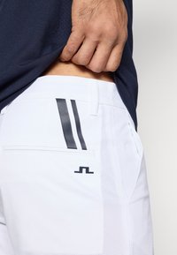 Λευκά αθλητικά σόρτς με μαύρες λεπτομέρειες. Διαθέτουν δύο κάθετες μαύρες ρίγες και ένα μικρό λογότυπ ο στην πίσω τσέπη. Ελαφρύ, υφασμάτινο υλικό.