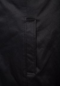 Svart tyg med en slät textur har en vertikal söm och en rektangulär insatsficka. Stygnarna är precisa och enhetliga.