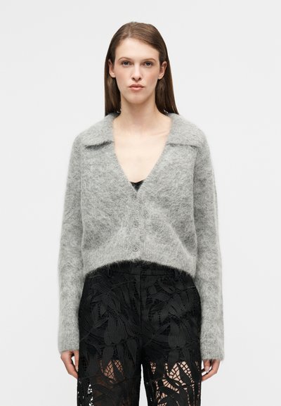 Ανοιχτό γκρι, χνουδωτό ζακέτα με γιακά και κούμπωμα με κουμπιά, συνδυασμένο με περίτεχνες μαύρες δαντελένιες παντελόνες με φύλλο μοτίβο.