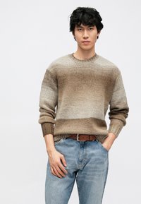 Stickad tröja med ett gradientmönster i beige och bruna toner, med rund hals och ribbade muddar, kombinerad med ljusblå jeans och ett brunt bälte.