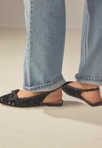 Chaussures slingback noires pailletées avec un détail de nœud en satin, semelle plate et talon ouvert, associées à un jean large bleu clair.