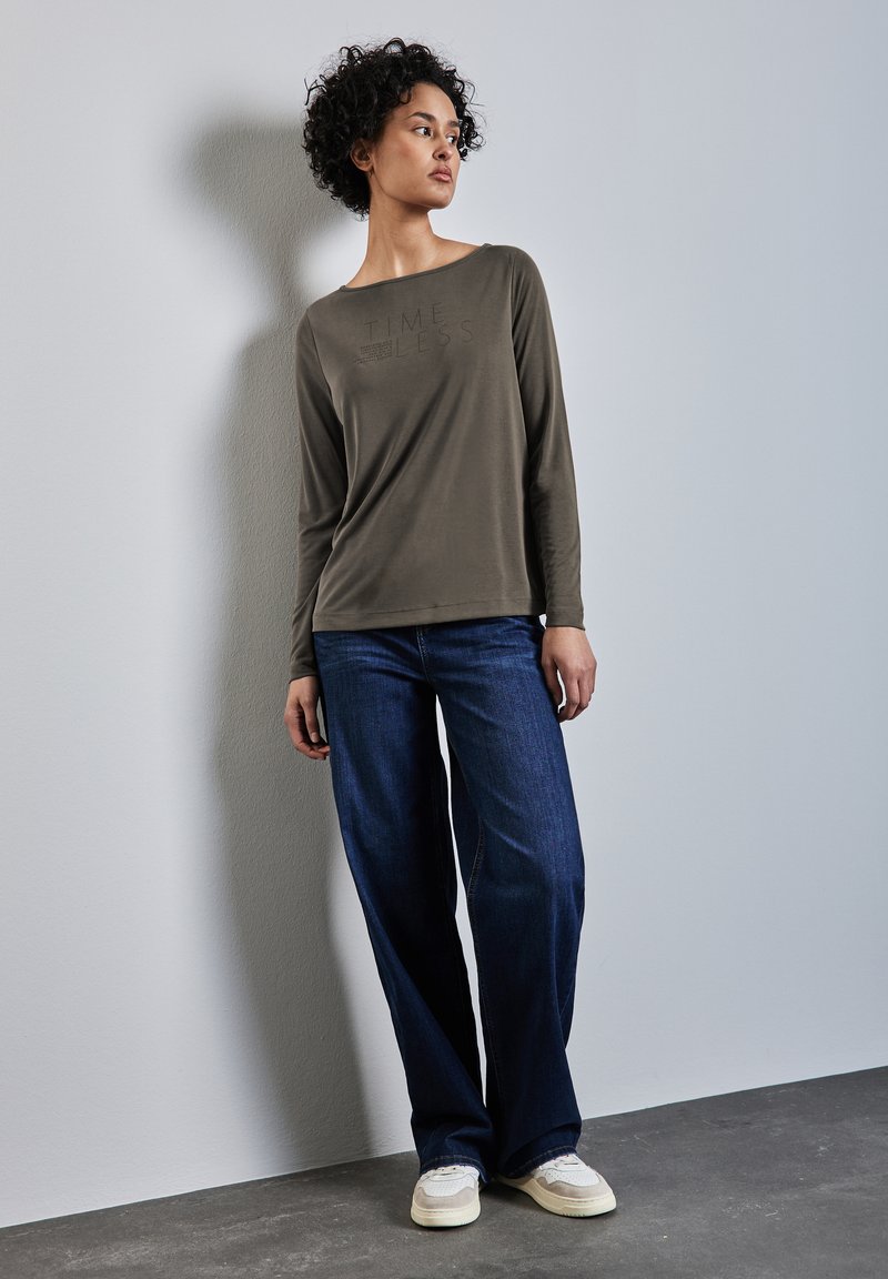 Maglia a maniche lunghe verde oliva con testo "TIME LESS", abbinata a jeans blu scuro larghi e sneakers chiare.