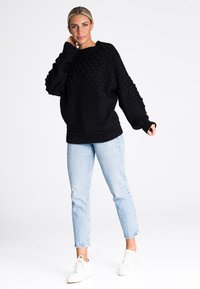 Czarny, oversize sweter dzianinowy z teksturowanym wzorem z przodu, zestawiony z jasnoniebieskimi spodniami o prostym kroju i białymi sneakersami.