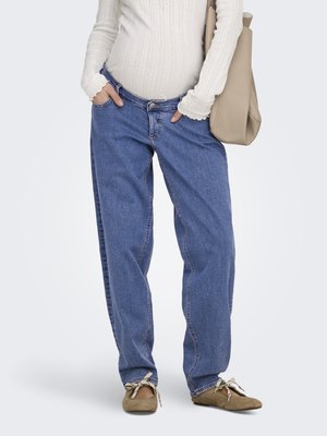 Osoba nosi voľné modré rifle, béžové balerínky s mašľami, biely rebrovaný dlhý rukáv a nesie béžovú tašku cez rameno.