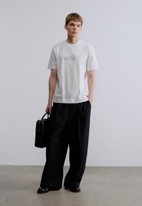 Λευκό t-shirt με λεπτό μπλε σχέδιο, συνδυασμένο με φαρδιά μαύρη παντελόνια. Το μοντέλο κρατά μαύρη τσάντα, στέκεται μπροστά σε ουδέτερο φόντο.