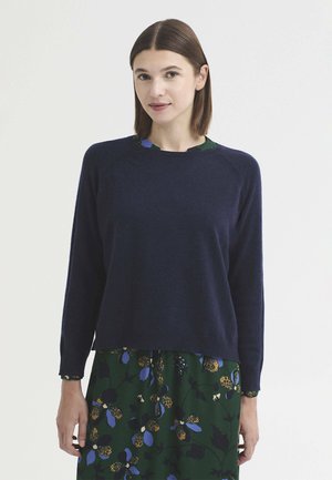 Femme aux cheveux bruns et raides portant un pull bleu marine par-dessus une robe verte avec des motifs floraux bleus et bruns sur un fond uni.