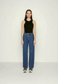 Top preto sem mangas combinado com jeans azuis de perna larga e cintura alta. A modelo usa ténis pretos e brancos. Design de roupa simples e casual.