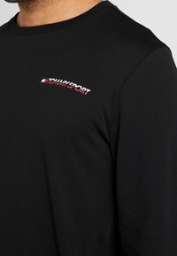 Černá dlouhá bavlněná košile s malým červeným a bílým logem "TOMMY SPORT" na hrudi. Kulatý výstřih a přiléhavý střih.