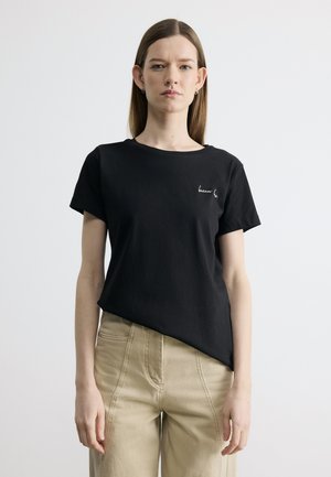 Žena s rovnými hnedými vlasmi, oblečená v čiernom tričku s krátkym rukávom a béžových nohaviciach, stojaca pred jednoduchým svetlým pozadím.