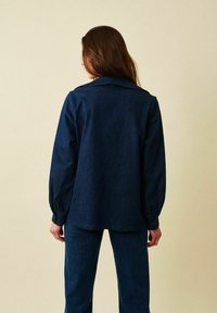 Mörkblå jeansskjorta med krage, lång ärm och knappmanschetter. Slät textur, lös passform och rak fåll. Matchas med byxor i samma färg.