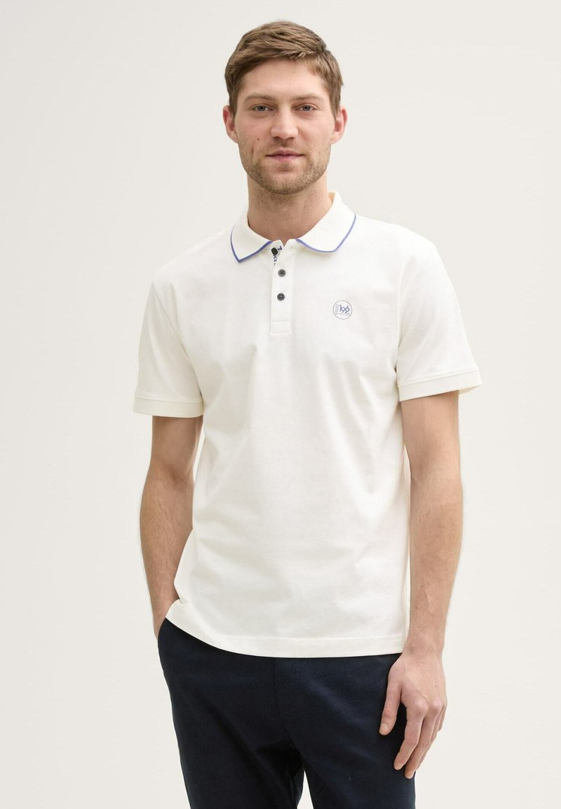 Polo shirt bianco in cotone con colletto a punta, rifiniture blu, patta con tre bottoni e un piccolo logo sul lato sinistro del petto. Maniche corte e vestibilità regolare.