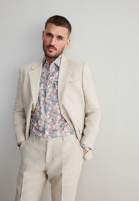 Beige linnen pak met een gestructureerde revers, gecombineerd met een licht bloemenpatroon overhemd in pastelkleuren. Model staat met handen in de zakken.