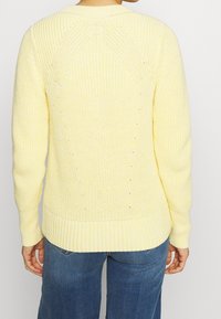 Gul stickad tröja med ribbad fåll och ärmar, med en strukturerad yta och en lös passform. Baksidan visar sömdetaljer.