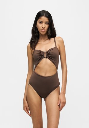 Femme aux longs cheveux foncés portant un maillot de bain une pièce marron avec un buste froncé et une découpe sur le devant, sur un fond clair uni.