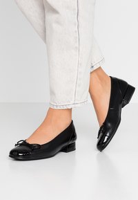 Black patent leather ballet flats with a rounded toe, small heel, and decorative bow on the front, paired with light-colored jeans.