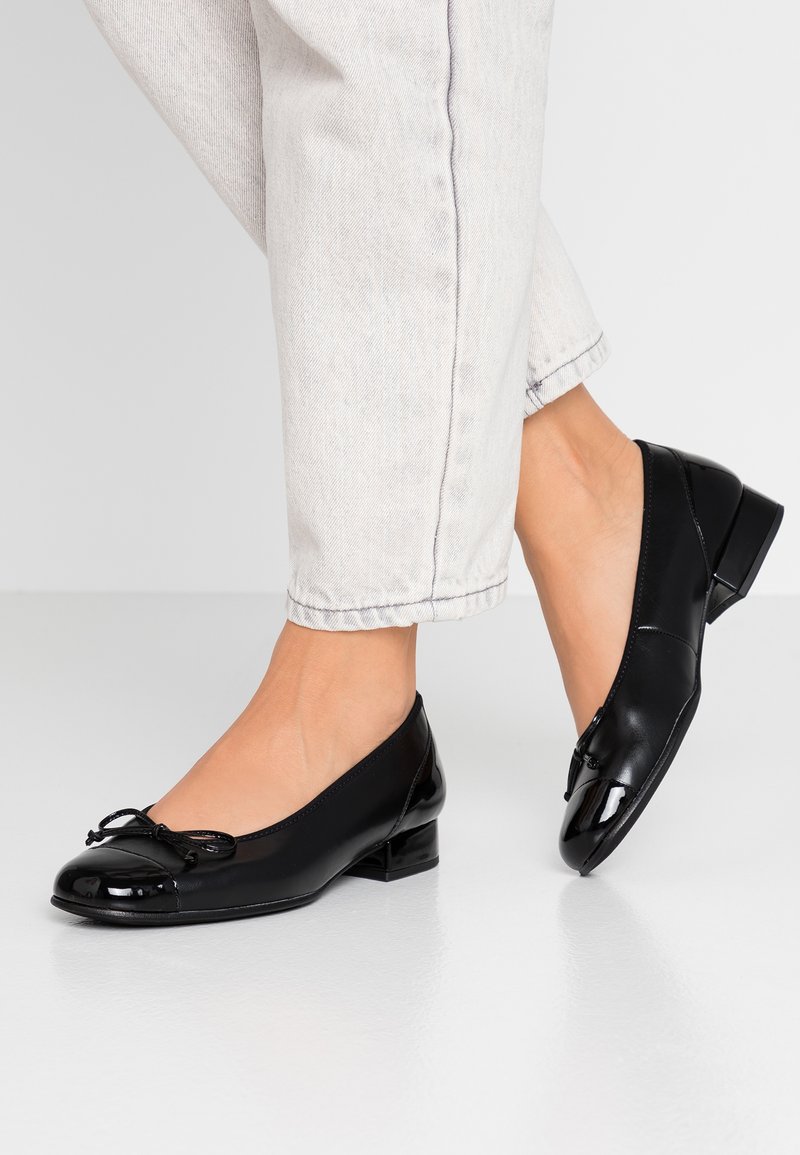 Black patent leather ballet flats with a rounded toe, small heel, and decorative bow on the front, paired with light-colored jeans.
