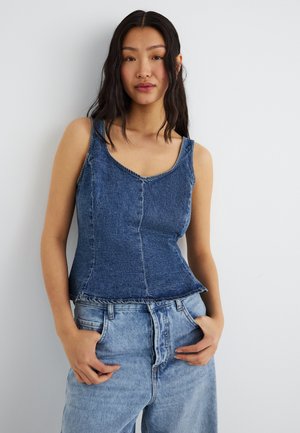 Mladá žena s dlhými tmavými vlasmi, oblečená v priliehavom modrom riflovom tielku bez rukávov a svetlomodrých rifle, stojaca s rukami v kapsách.