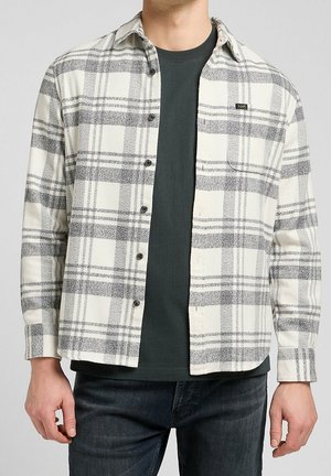 Mann trägt ein cremeweiß-grau kariertes Hemd über einem dunkelgrauen Rundhalsausschnitt-T-Shirt und dunkler Jeans.